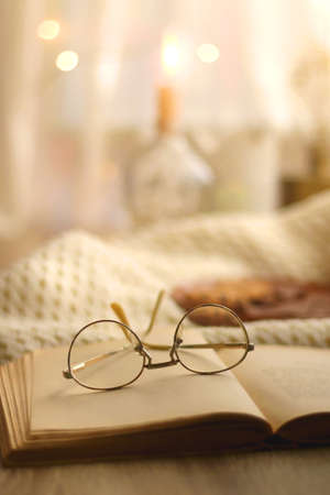 Open book, reading glasses, soft blanket and plate with chocolate and chocolate chip cookies. Hygge at home. Selective focus.の写真素材