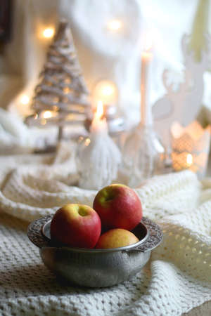 Bowl of apples and soft blanket with lit candles and Christmas decorations in the background. Selective focus.の写真素材