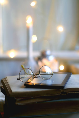 Stack of vintage boooks, digital e-reader, reading glasses, cozy blanket and lit candles. Hygge at home. Selective focus.の写真素材