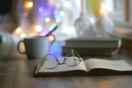 Cup of warm drink with cinnamon stick and candy cane, open book, reading glasses, lit candles and colorful bokeh lights. Selective focus.の写真素材