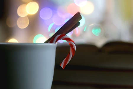 Cup of warm drink with cinnamon stick and candy cane, open book, reading glasses, lit candles and colorful bokeh lights. Selective focus.の写真素材