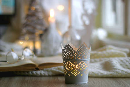Open book and reading glasses on the table. Lit candles, soft blanket and Christmas decorations in the background. Hygge at home. Selective focus.の写真素材