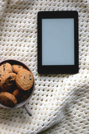 Digital e-reader or tablet, bowl of chocolate chip cookies and soft knitted blanket on wooden table. Hygge at home. Flat lay.の写真素材