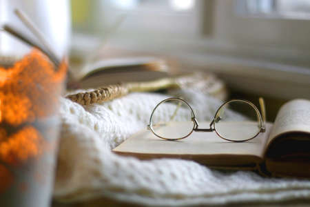 Candle holder with lit candle, open book and reading glasses, soft knitted blanket and wicker basket. Hygge at home. Selective focus.の写真素材