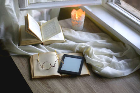 Open books, reading glasses, e-reader device and lit candle on the table. Cozy reading at home....
