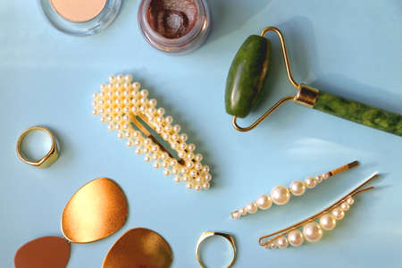 Various make-up products, jade face roller, pearl hair accessories, earrings and gold rings on bright blue background. Flat lay.の写真素材