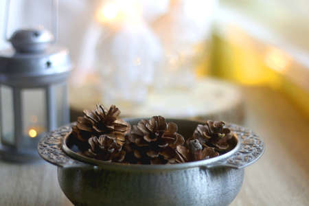 Silver bowl filled with natural pinecones, lantern and lit candles on the table. Hygge at home....