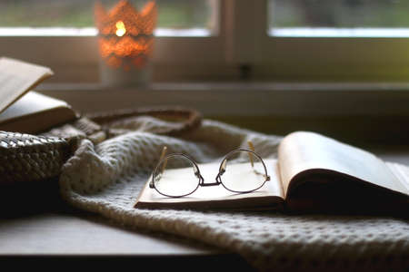 Candle holder with lit candle, open book and reading glasses, soft knitted blanket and wicker basket. Hygge at home. Selective focus.の写真素材