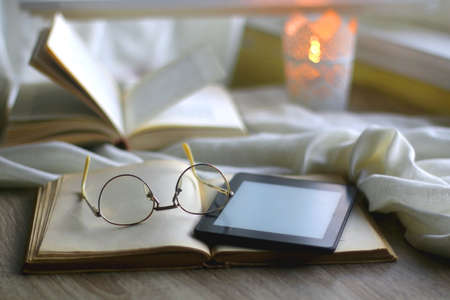 Open books, reading glasses, e-reader device and lit candle on the table. Cozy reading at home. Selective focus.の写真素材