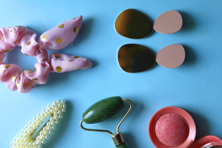 Jade roller, pink blush, fashionable earrigns, pink scrunchie and pearl barrette on bright blue background. Flat lay.の写真素材