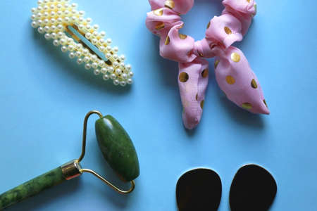 Jade roller, pink blush, fashionable earrigns, pink scrunchie and pearl barrette on bright blue background. Flat lay.の写真素材