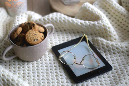 Bowl of chocolate chip cookies, digital tablet, reading glasses, soft blanket and lit candle. Hygge...