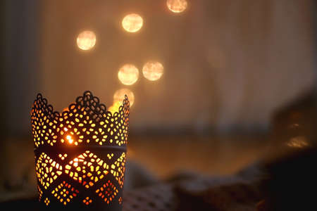 Candle holder with lit candle, open book, reading glasses and soft blanket. Bokeh lights in the...