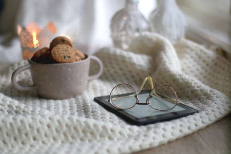 Bowl of chocolate chip cookies, digital tablet, reading glasses, soft blanket and lit candle. Hygge...
