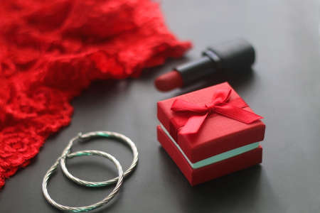 Small red giftbox, red lipstick, silver earrings and lace underpants. Valentine's Day objects on dark background. Selective focus.の写真素材