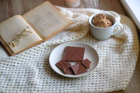 Plate with almond chocolate bar, mug with chocolate chip cookies, open book, reading glasses, soft...
