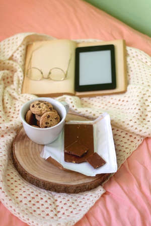 Mug of chocolate chip cookies, bar of chocolate, open book, reading glasses and e-reader on a bed. Selective focus.の写真素材