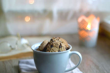 Cup of chocolate chip cookies, open book and reading glasses on the table. Lit candle and bokeh...