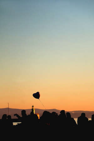 Silhouette of unrecognizable group of people and heart shaped balloon, enjoying sunset. Selective focus.の写真素材