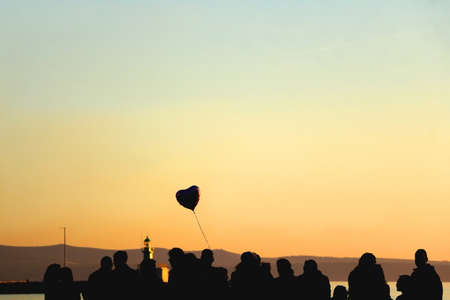 Silhouette of unrecognizable group of people and heart shaped balloon, enjoying sunset. Selective focus.の写真素材