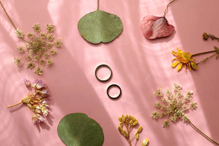 Gold wedding rings and various pressed flowers on pastel pink background, illuminated by sunlight. Flat lay.の写真素材