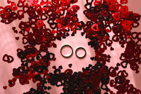 Gold wedding rings and red heart confetti on pastel pink background. Flat lay.