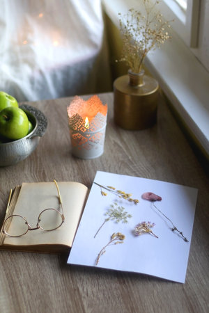 Open book, reading glasses, paper with pressed flowers, bowl of apples and lit candle on the table. Hygge at home. Selective focus.の写真素材