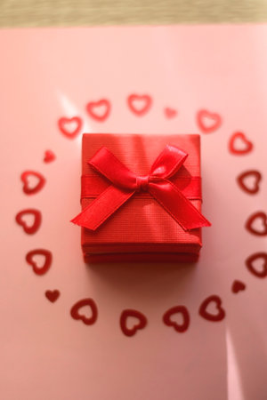 Small red giftbox and heart confetti on pale pink background. Selective focus.