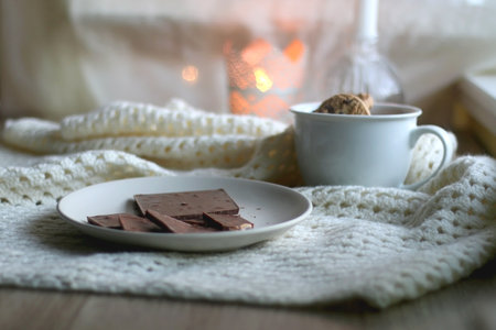 Plate with almond chocolate bar, mug with chocolate chip cookies, open book, reading glasses, soft...