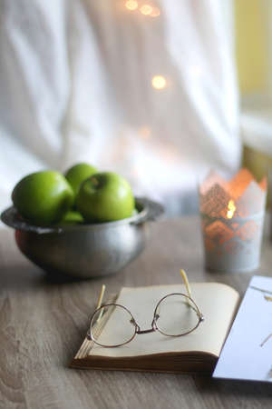 Open book, reading glasses, paper with pressed flowers, bowl of apples and lit candle on the table. Hygge at home. Selective focus.の写真素材