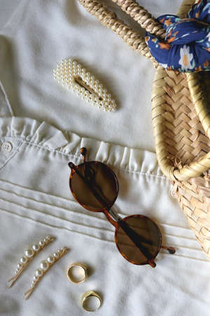 White blouse, wicker bag with tied floral scarf, tortoiseshell sunglasses, pearl hair clips and gold rings. Fashion for spring and summer. Flat lay.の写真素材