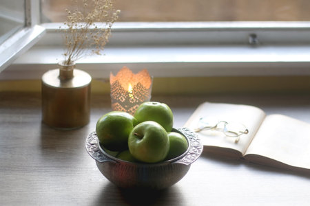 Bowl of green apples, open book, reading glasses, lit candle and flowers on the table. Selective focus.の写真素材