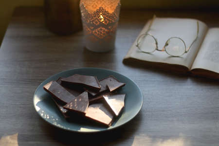 Plate of chocolate, open book, reading glasses, lit candle and flowers on the table. Selective...