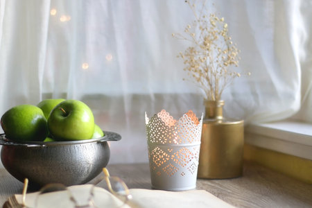 Bowl of green apples, open book, reading glasses, lit candle and flowers on the table. Selective...