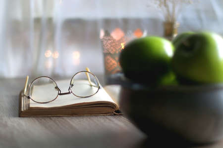 Bowl of green apples, open book, reading glasses, lit candle and flowers on the table. Selective focus.の写真素材
