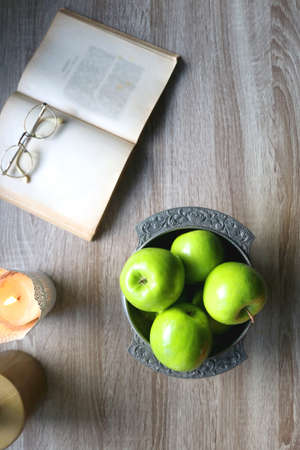 Bowl of green apples, open book, reading glasses, lit candle and flowers on the table. Top view.の写真素材