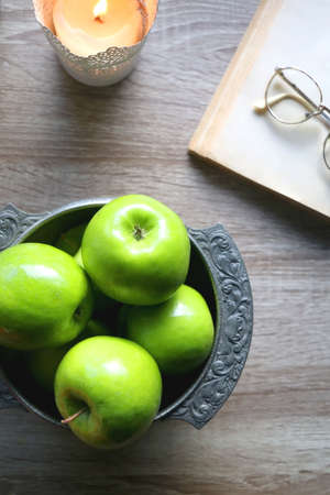 Bowl of green apples, open book, reading glasses, lit candle and flowers on the table. Top view.の写真素材