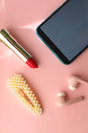 Mobile phone, pink wireless earphones, red lipstick and pearl hair clip on pastel pink background. Flat lay.の写真素材