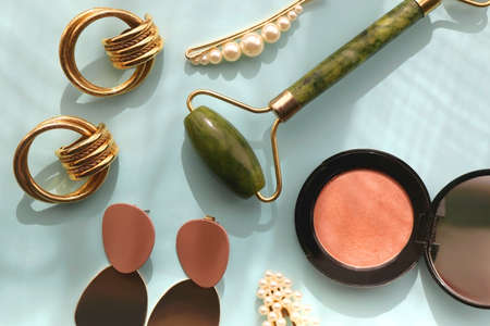 Coral blush, jade roller, pearl barrettes and gold earrings on bright blue background. Flat lay.の写真素材