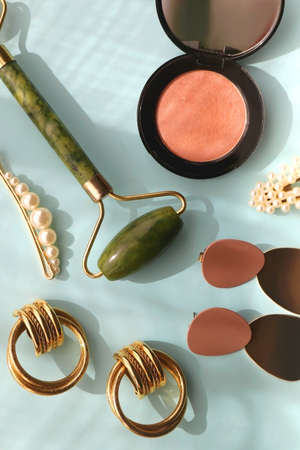 Coral blush, jade roller, pearl barrettes and gold earrings on bright blue background. Flat lay.