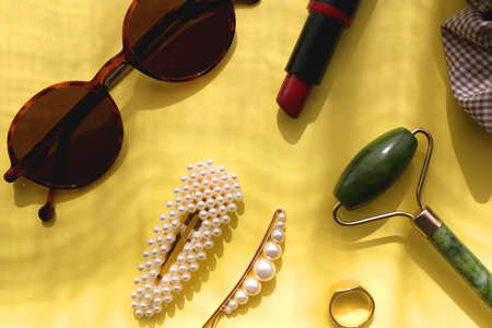 Round sunglasses, lipstick, jade roller, ring and hair accessories on bright yellow background. Flat...