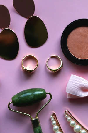 Pink scrunchie, gold earrings and rings, blush, various hair barrettes, lipstick and jade roller on bright pink background. Flat lay.の写真素材