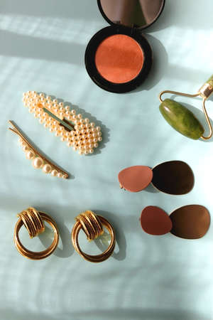 Coral blush, jade roller, pearl barrettes and gold earrings on bright blue background. Flat lay.の写真素材