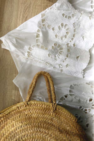 White linen lace blouse and round straw bag on wooden background. Flat lay.