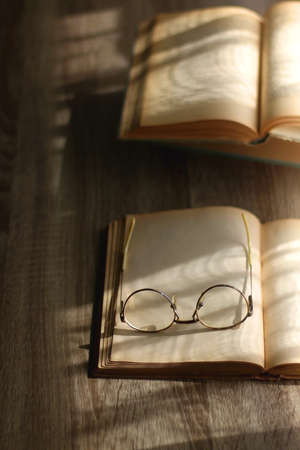 Open book and reading glasses on the table, illuminated by sunlight. Stack of vintage books in the background. Selective focus.の写真素材