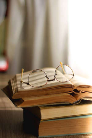 Open book and reading glasses on the table, illuminated by sunlight. Stack of vintage books in the background. Selective focus.の写真素材