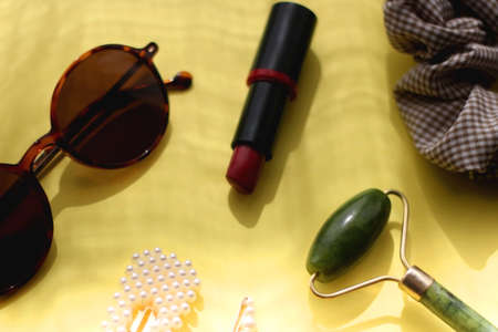 Round sunglasses, lipstick, jade roller, ring and hair accessories on bright yellow background....