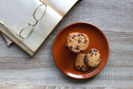 Plate of chocolate chip cookies, open book and reading glasses on wooden table. Flat lay.の写真素材