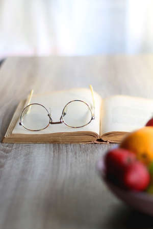 Bowl of apples, oranges and strawberries, open book and reading glasses on the table. Selective focus.の写真素材