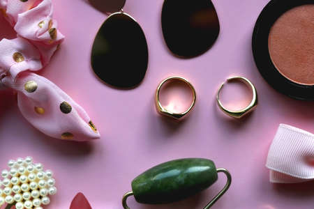 Pink scrunchie, gold earrings and rings, blush, various hair barrettes, lipstick and jade roller on bright pink background. Flat lay.の写真素材